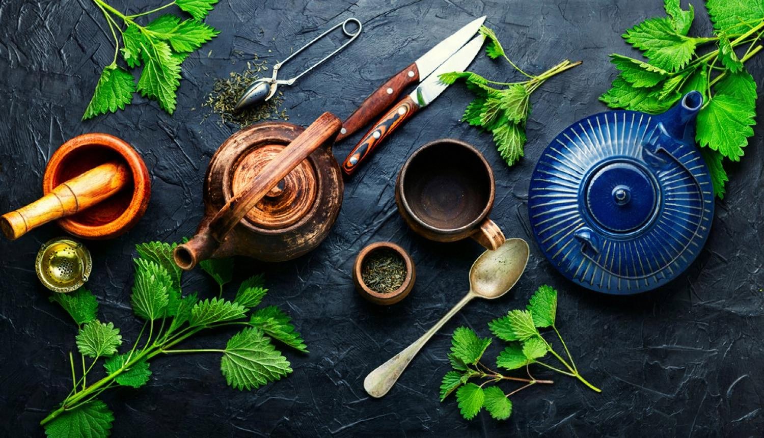 Overhead view of green tea brewing equipment, strainer, teapot and fresh leaves on a dark surface