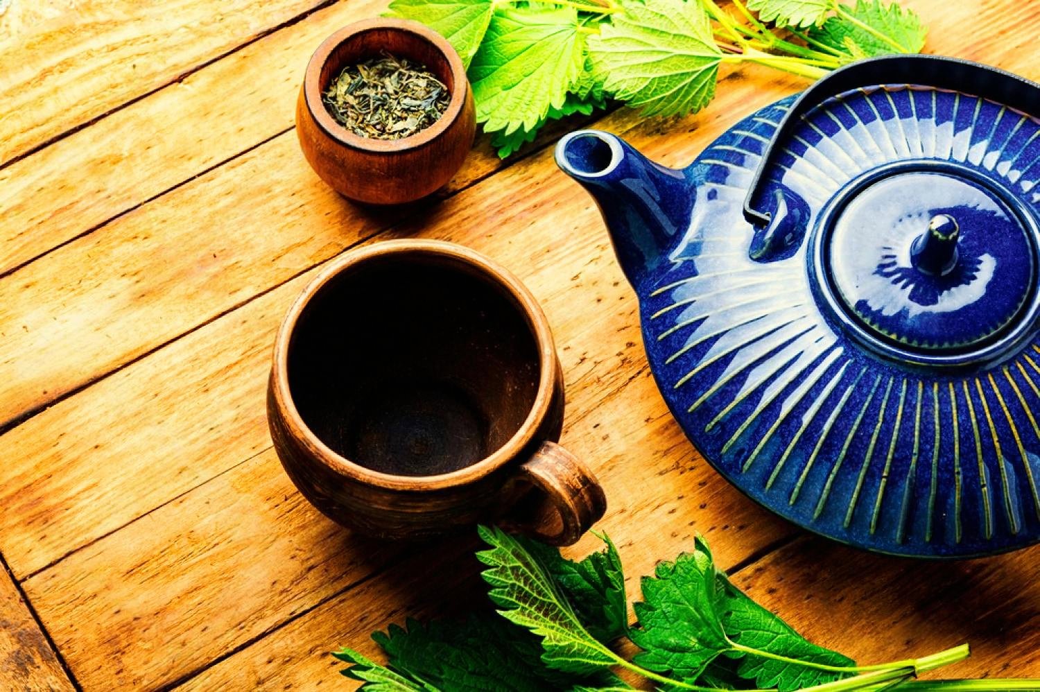 Loose green tea leaves in a wooden bowl next to a blue Japanese teapot and clay cup